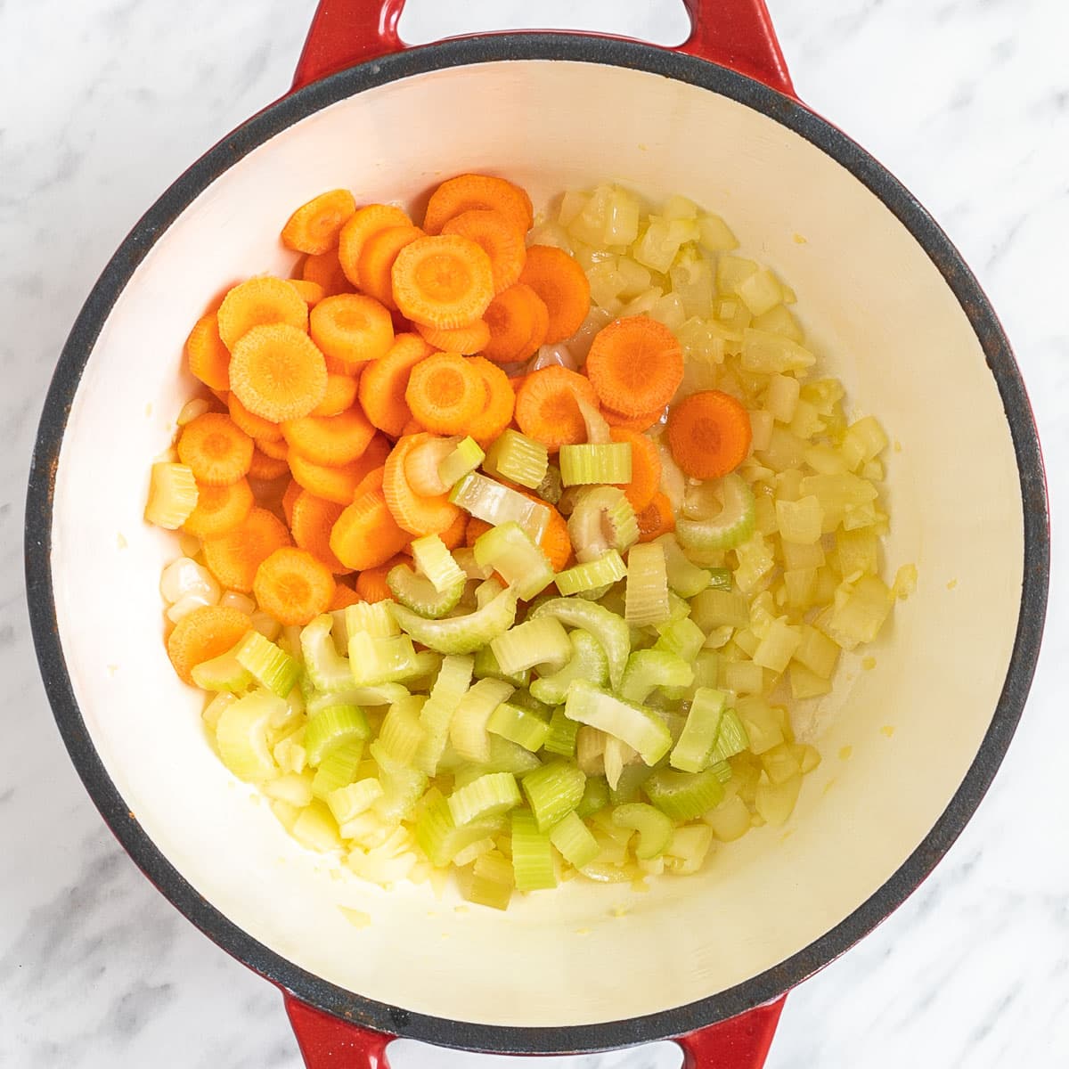 A red-white enameled Dutch oven with chopped veggies like carrot, celery and onion.