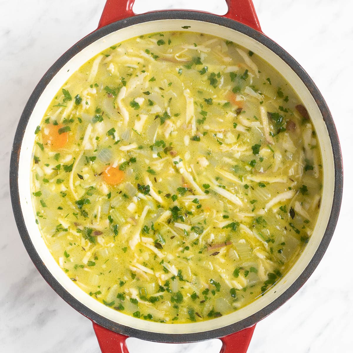 A red-white enameled Dutch oven with chopped veggies like carrot, celery, and onion, shredded mushroom pieces, green herbs in thick white stew.