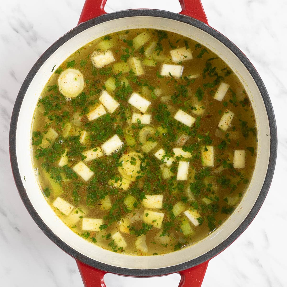 Red white enameled Dutch oven with chopped veggies and green herbs in a light vegetable broth.
