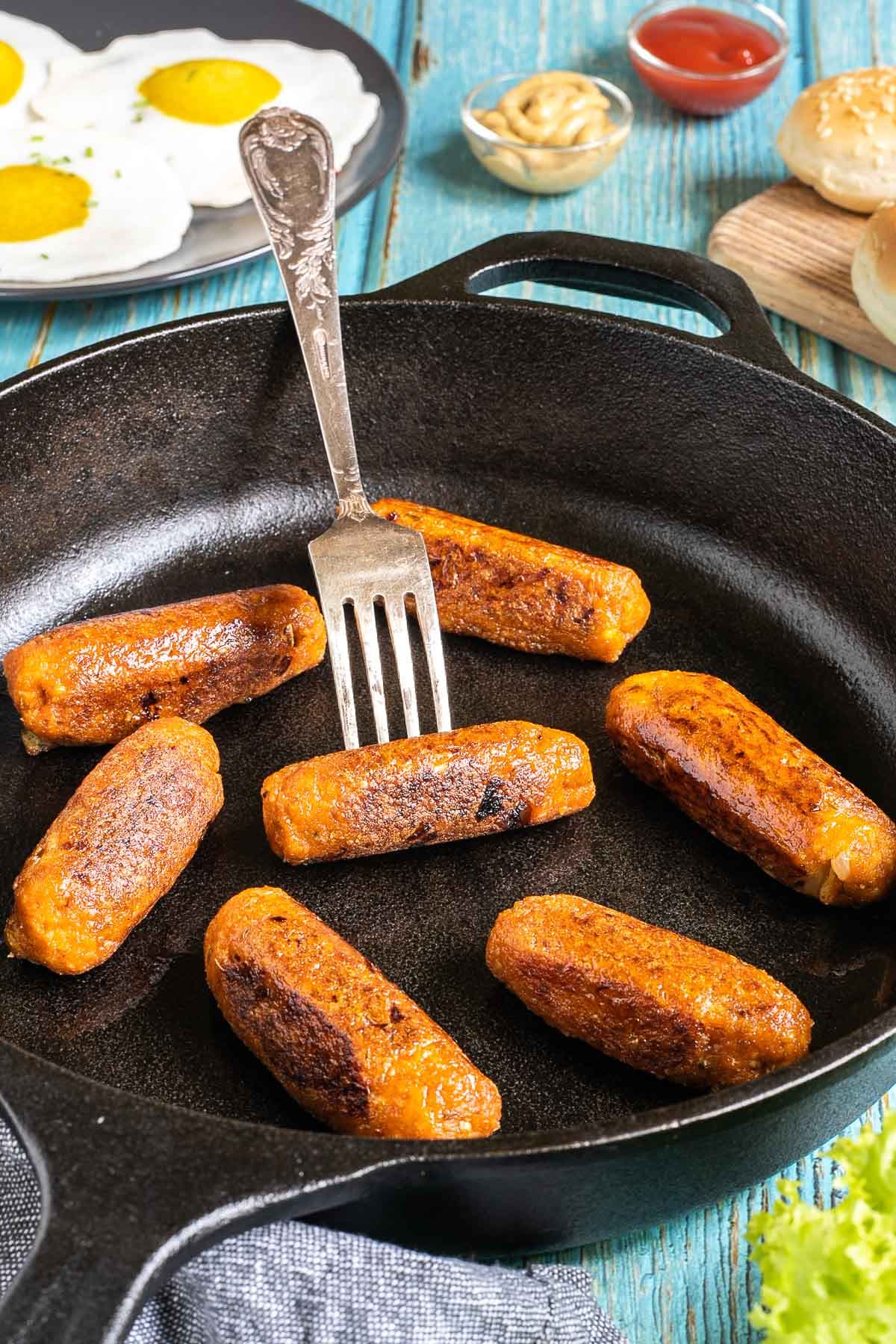 7 small sausages in a black cast-iron skillet. A Fork is pierced in one of them. Fake fried eggs, hoagie rolls, ketchup, and mustard are around it.