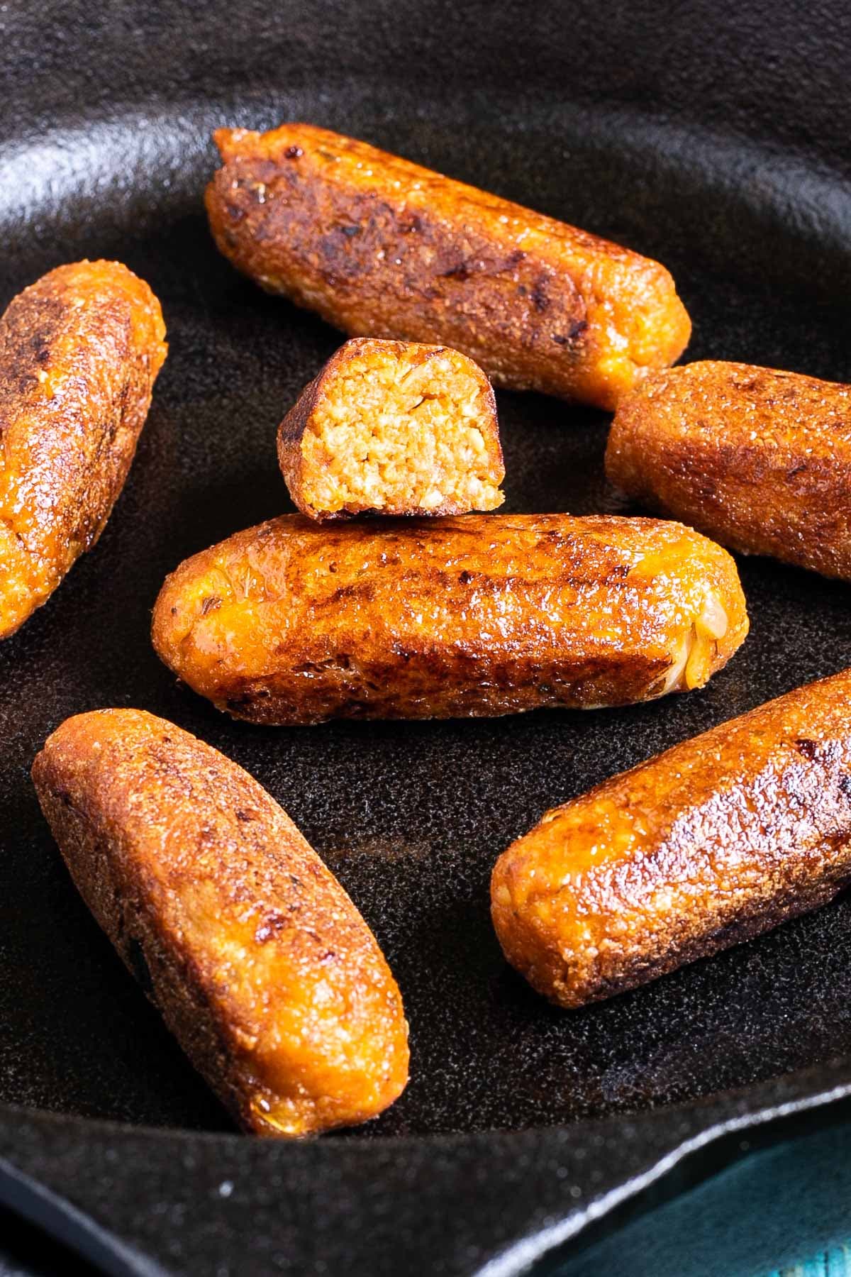 7 small sausages in a black cast-iron skillet. One is cut in half so the inside is visible.