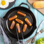 7 small sausages in a black cast-iron skillet. A hand holding tongs is taking one. Fake fried eggs, hoagie rolls, ketchup, and mustard are around it.
