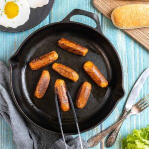 7 small sausages in a black cast-iron skillet. A hand holding tongs is taking one. Fake fried eggs, hoagie rolls, ketchup, and mustard are around it.