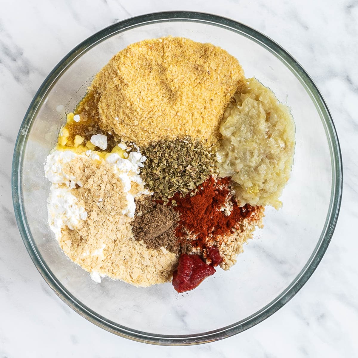 All ingredients for vegan sausage is in a glass bowl before mixing them. All ingredients are visible, tofu mince, breadcrumbs, starch, different spices and onion-garlic puree