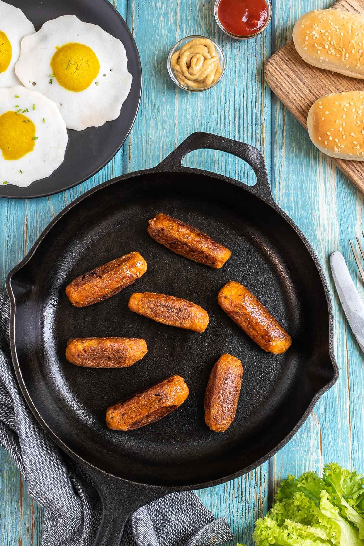 7 small sausages in a black cast-iron skillet. Fake fried eggs, hoagie rolls, ketchup, and mustard are around it.