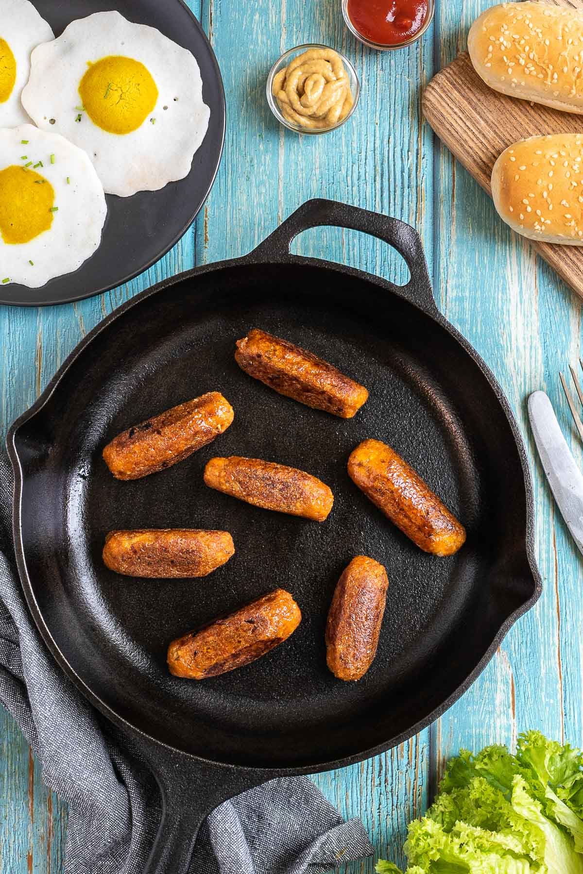 7 small sausages in a black cast-iron skillet. Fake fried eggs, hoagie rolls, ketchup, and mustard are around it.