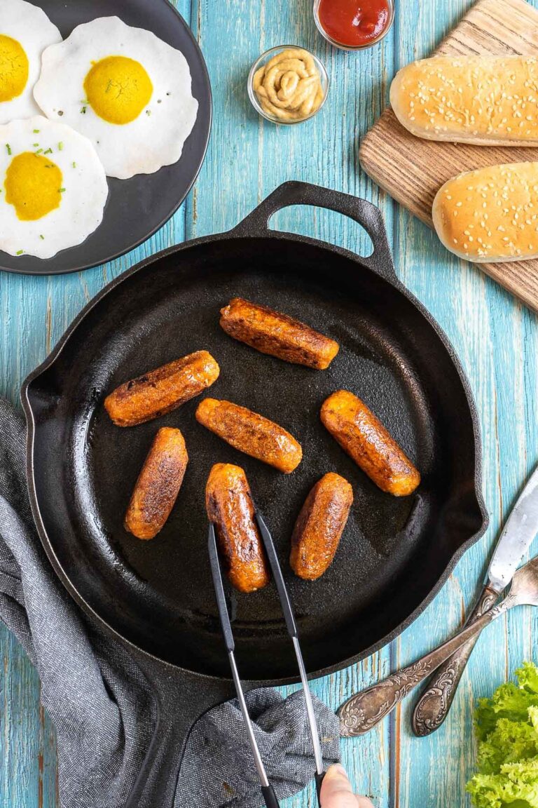 7 small sausages in a black cast-iron skillet. A hand holding tongs is taking one. Fake fried eggs, hoagie rolls, ketchup, and mustard are around it.