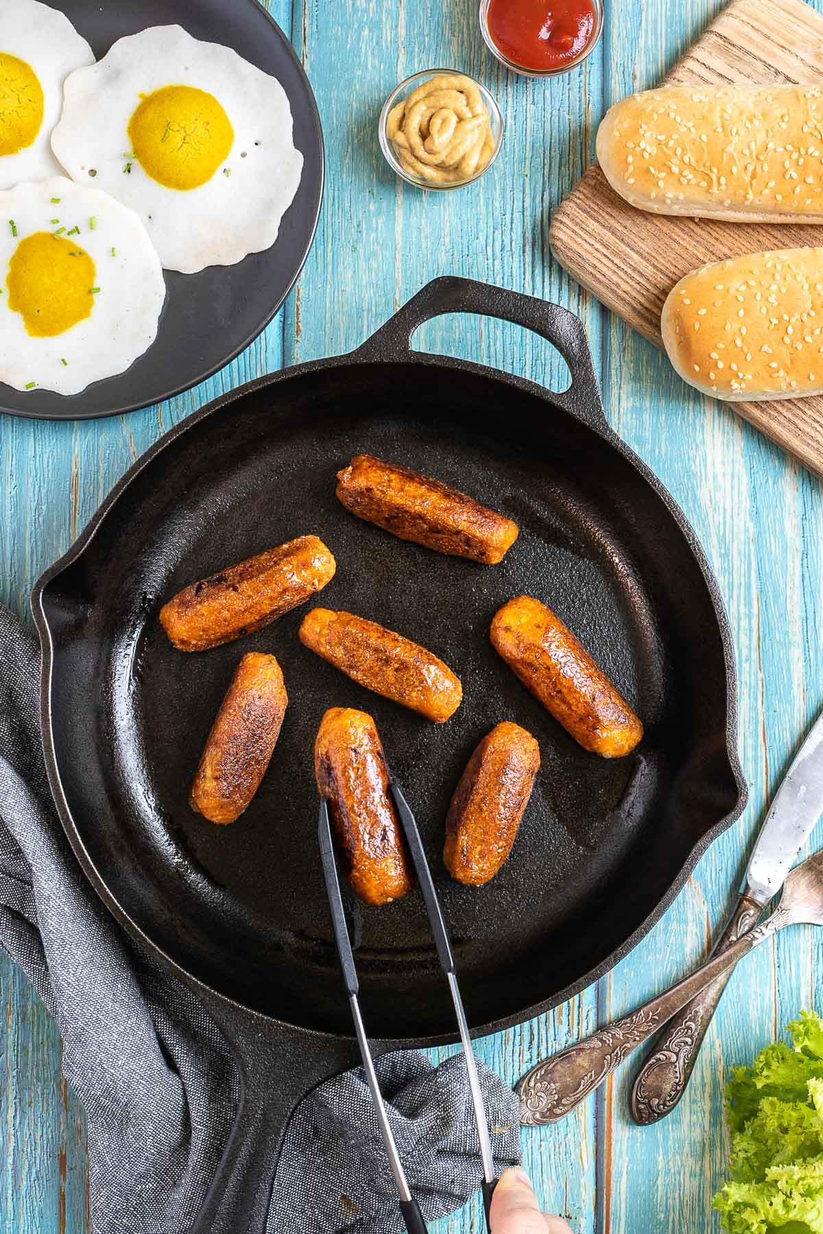 7 small sausages in a black cast-iron skillet. A hand holding tongs is taking one. Fake fried eggs, hoagie rolls, ketchup, and mustard are around it.