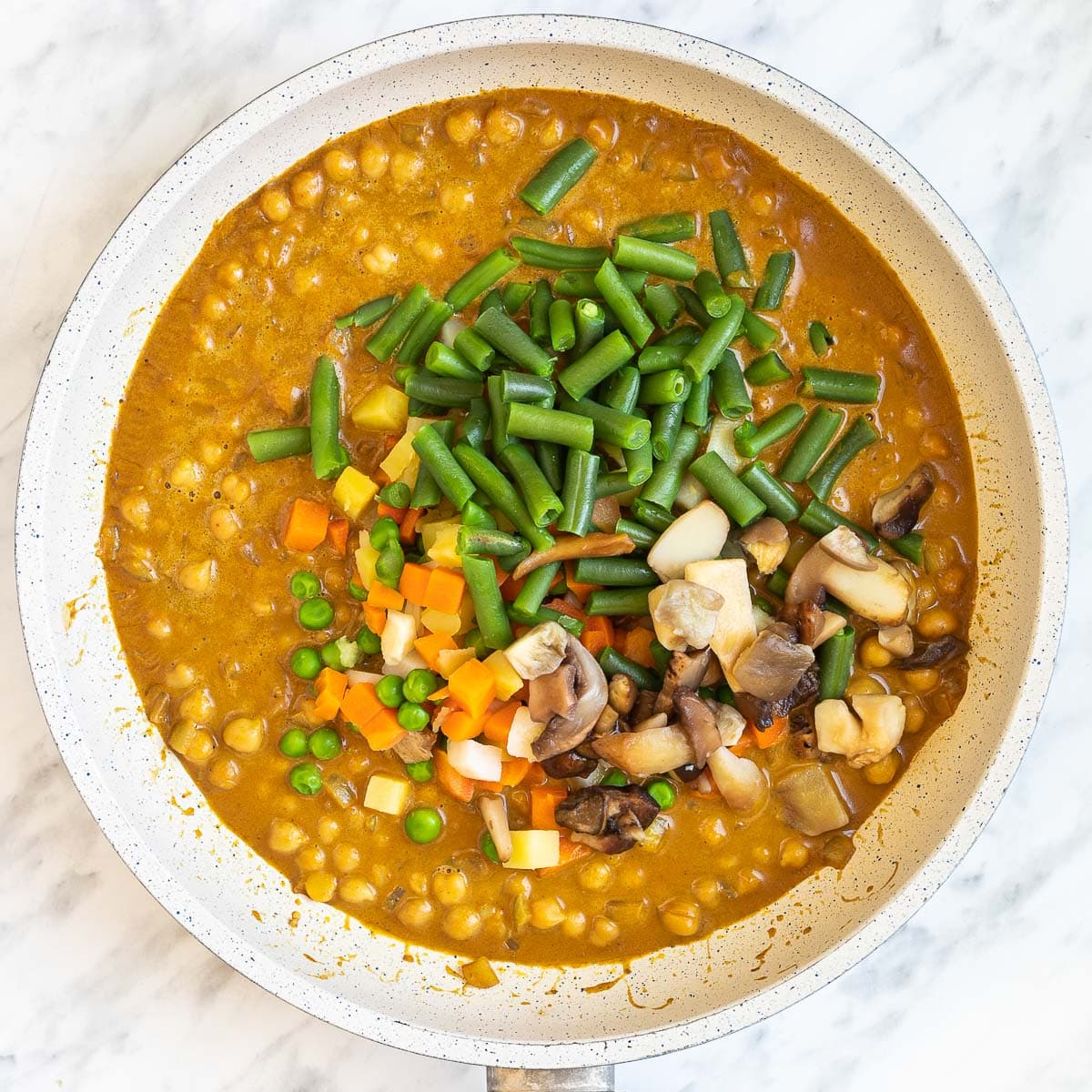 White frying pan with different chopped veggies added to the brown orange sauce like carrot, green bean, green peas, mushroom, chickpeas, and potato.