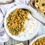 White plate with rice and different chopped vegetables, green peas, green beans, and chickpeas. Leftovers are next to it in the frying pan. Naan bread and more rice are on the side.