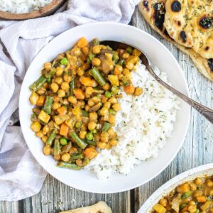 White plate with rice and different chopped vegetables, green peas, green beans, and chickpeas. Leftovers are next to it in the frying pan. Naan bread and more rice are on the side.