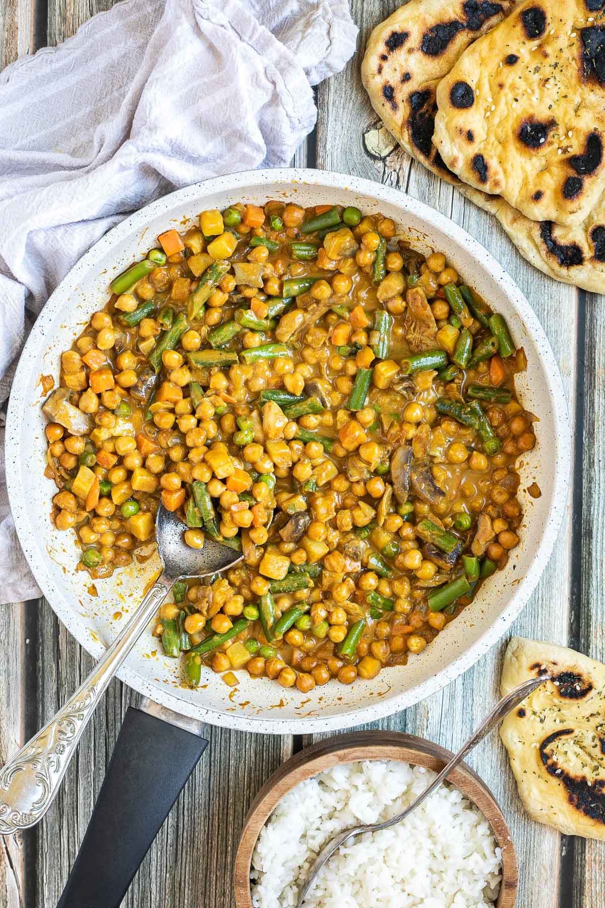 White frying pan full of vegetables like chickpeas, green beans, chopped carrots, green peas, chopped mushroom, and potatoes swimming in a brown-orange sauce. Naan bread and a brown bowl of rice are next to it.