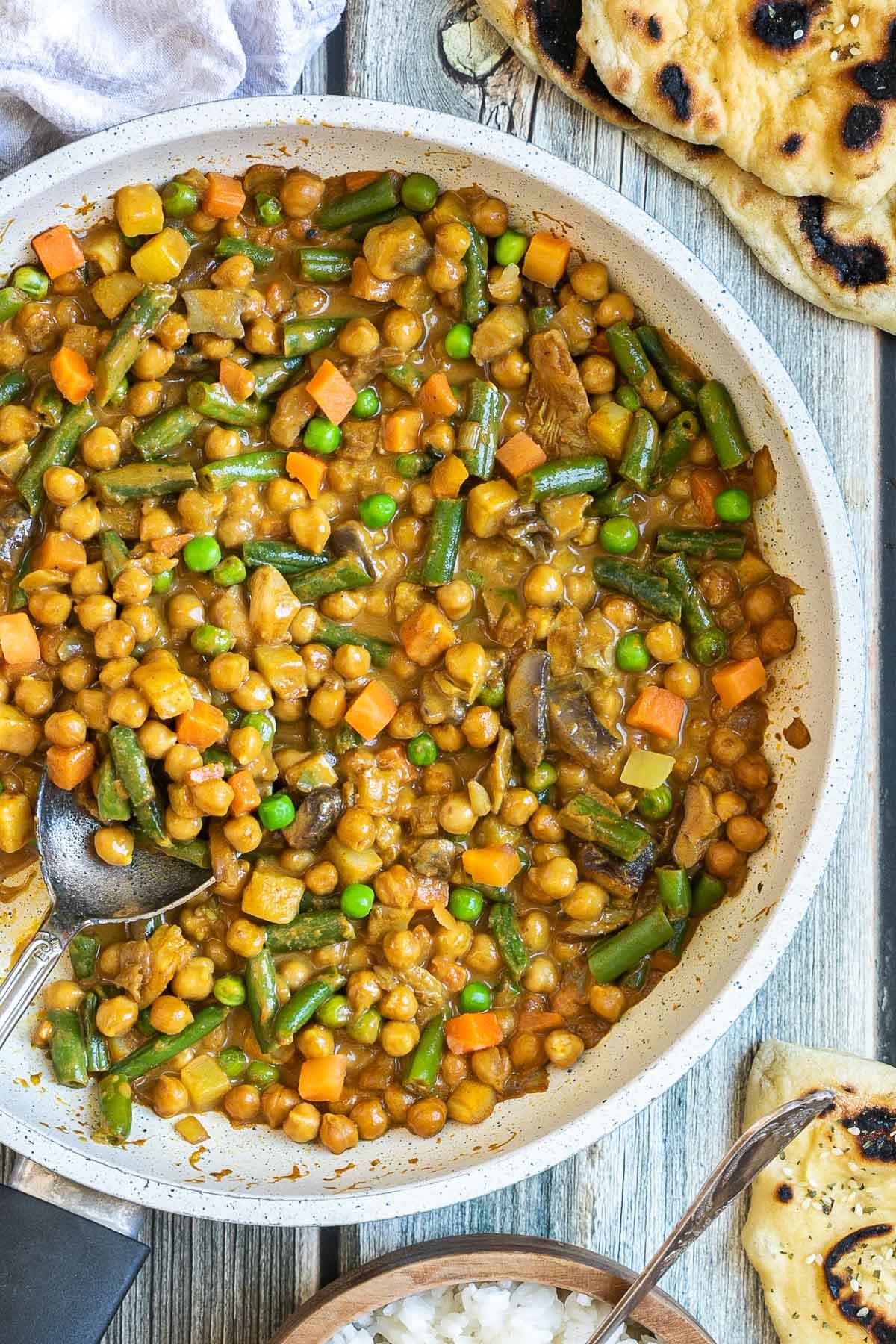White frying pan full of vegetables like chickpeas, green beans, chopped carrots, green peas, chopped mushroom, and potatoes swimming in a brown-orange sauce.