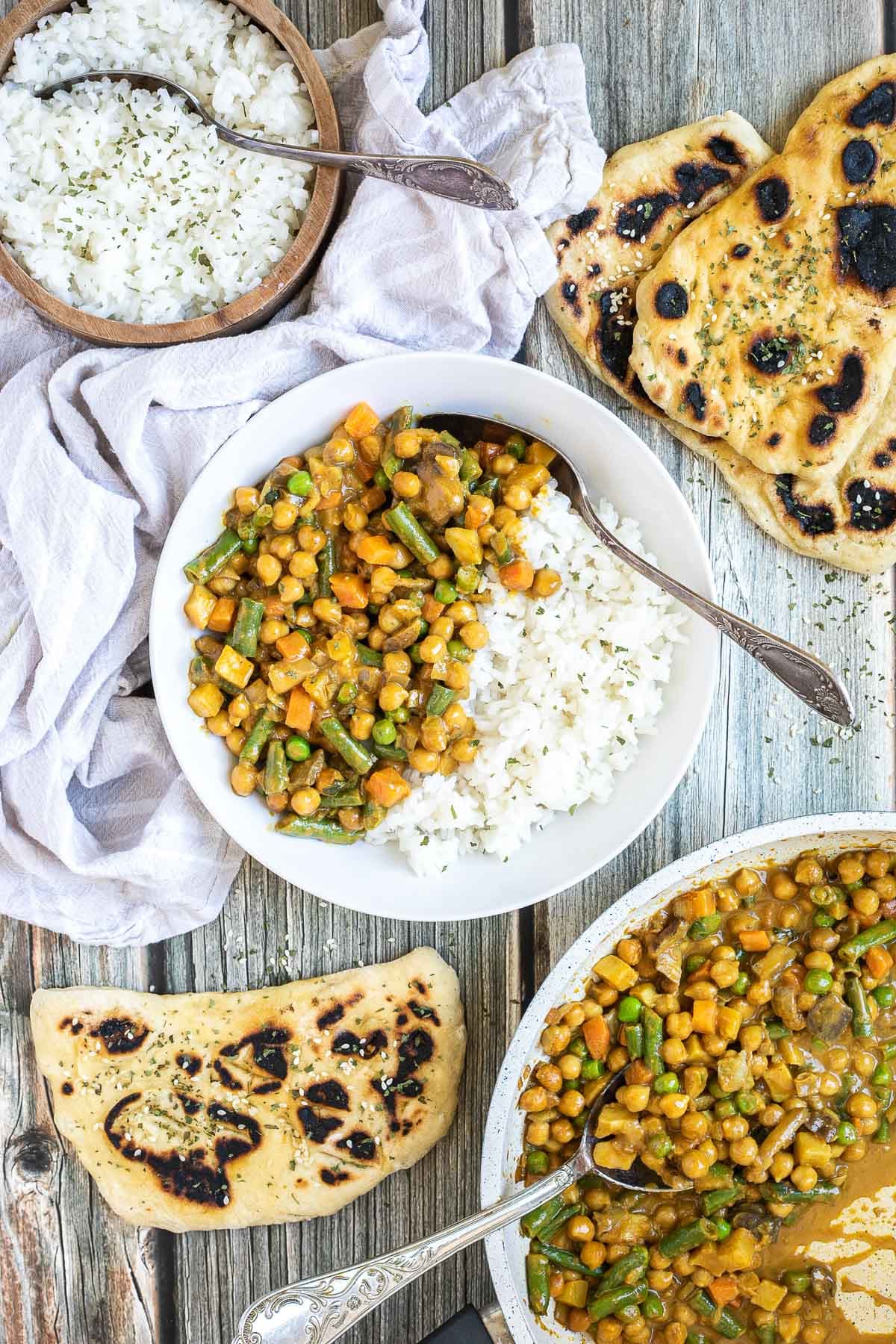 White plate with rice and different chopped vegetables, green peas, green beans, and chickpeas. Leftovers are next to it in the frying pan. Naan bread and more rice are on the side.