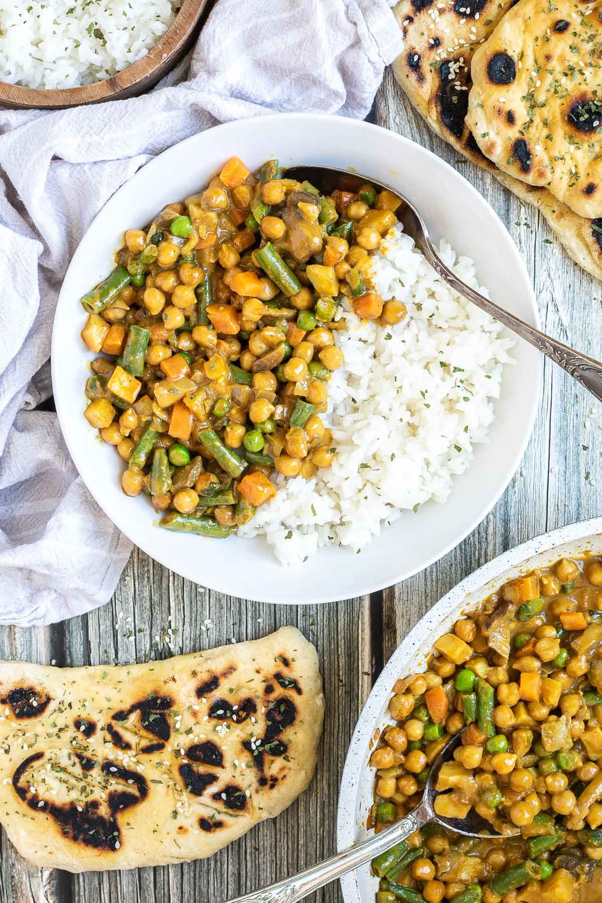 White plate with rice and different chopped vegetables, green peas, green beans, and chickpeas. Leftovers are next to it in the frying pan. Naan bread and more rice are on the side.
