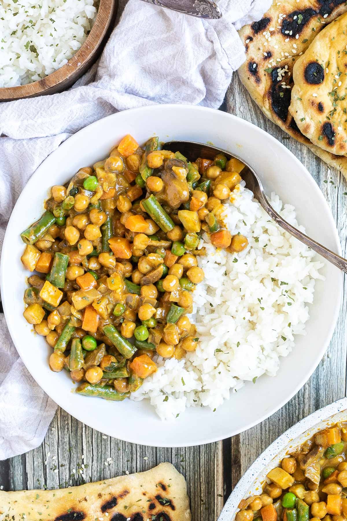 White plate with rice and different chopped vegetables, green peas, green beans, and chickpeas. Leftovers are next to it in the frying pan. Naan bread and more rice are on the side.