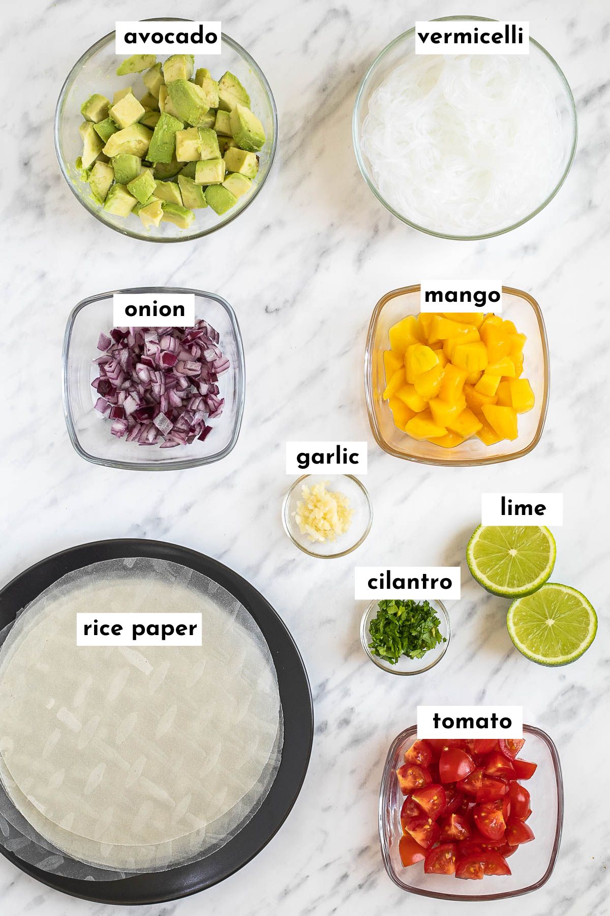 Ingredients of avocado spring rolls in small bowls like chopped avocado, chopped mango, chopped tomatoes, rice noodles, finely chopped red onion and garlic, chopped cilantro and lime. Rice papers are on a black plate.