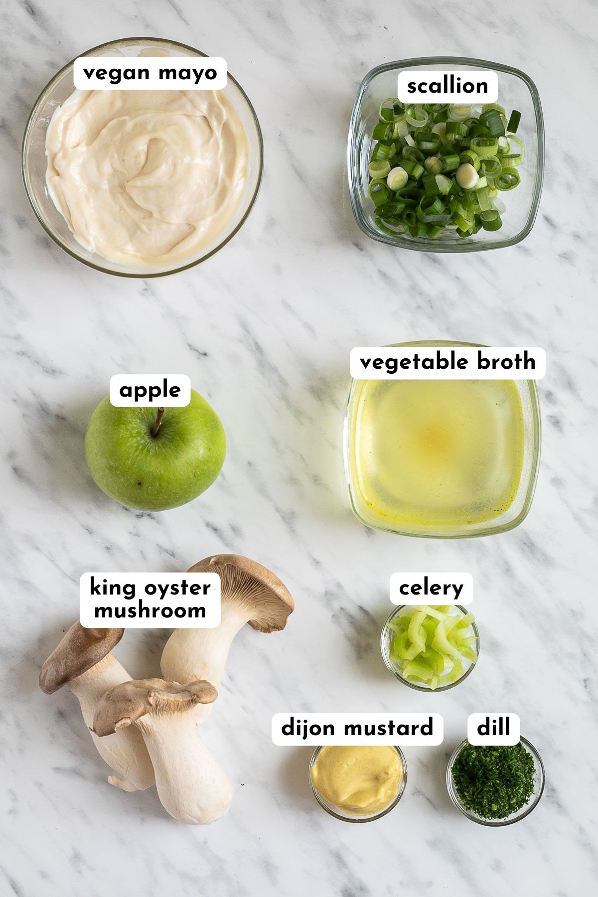 ingredients of vegan chicken salad on a marble surface bowl of mayo, bowl of broth, small bowl of mustard, king oyster mushrooms, a green apple, chopped scallion, dill and celery. 