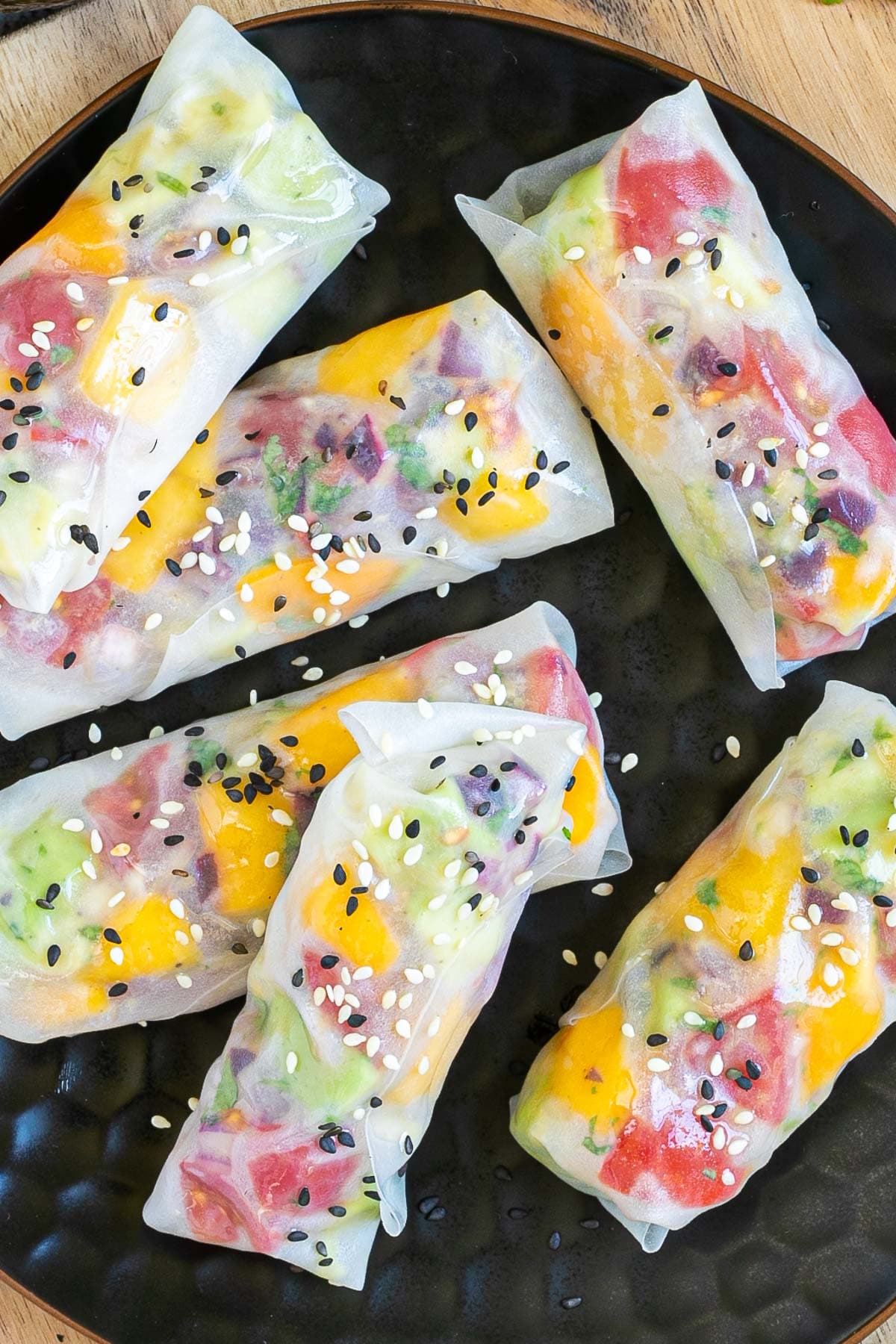 6 fresh rice paper wraps filled with chopped tomato, mango, and avocado placed on a black plate. Sprinkled with white and black sesame seeds.