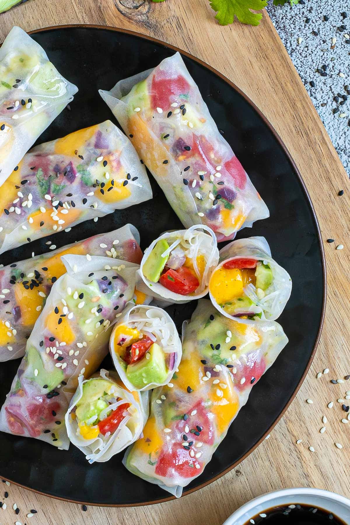 Fresh rice paper wraps filled with chopped tomato, mango, and avocado placed on a black plate. 4 half rolls and 6 whole rolls.