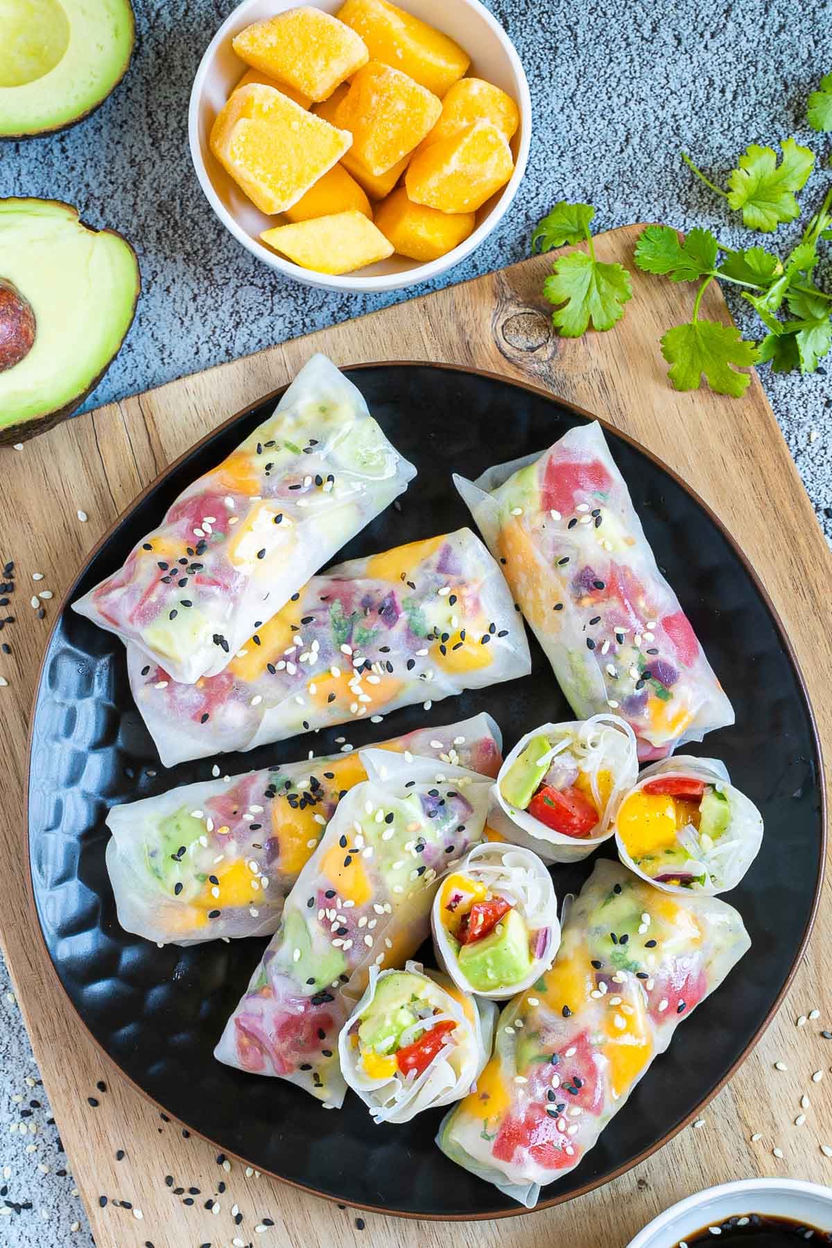 Fresh rice paper wraps filled with chopped tomato, mango, and avocado placed on a black plate. 4 half rolls and 6 whole rolls. Leftover ingredients are next to it. 