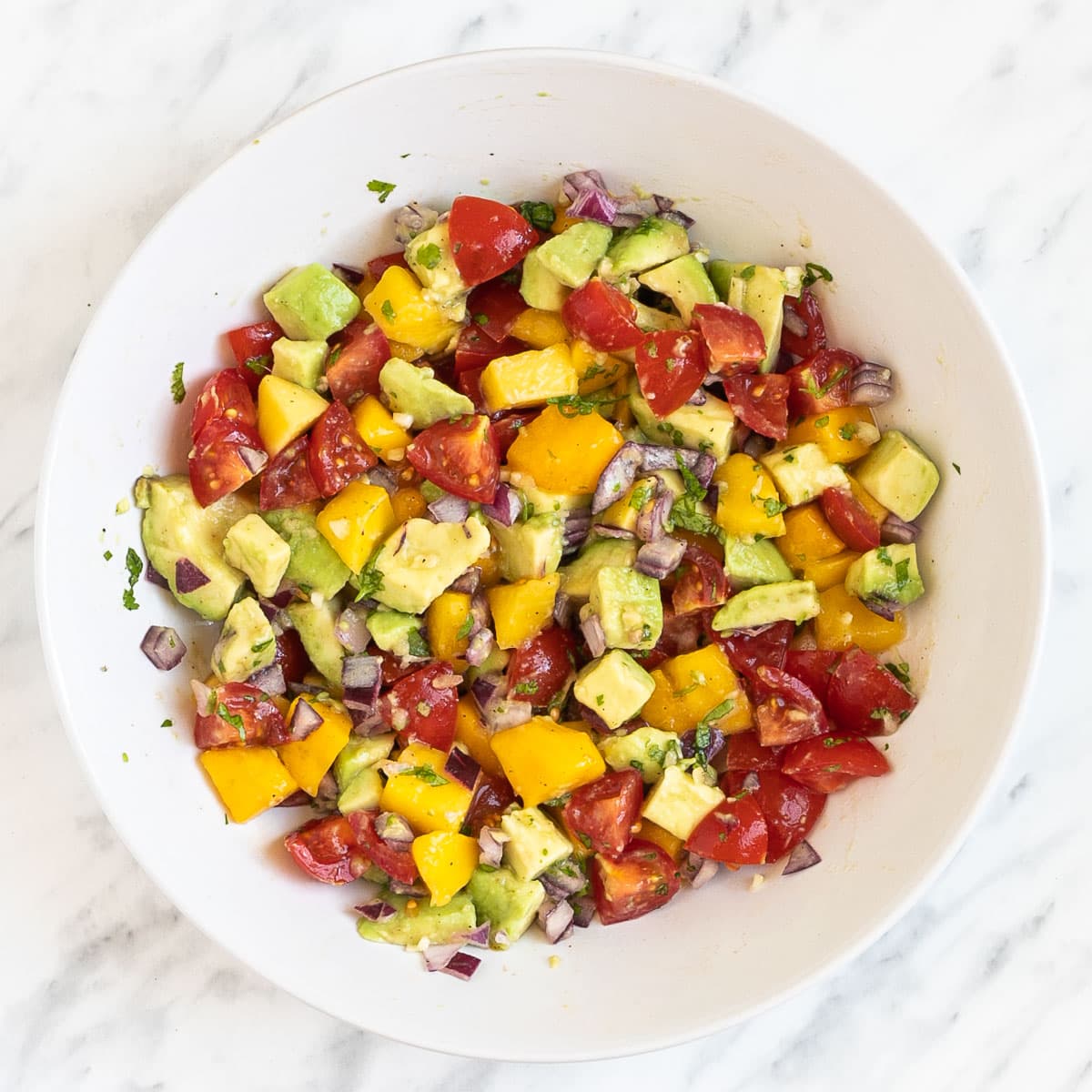 A white bowl of mixed chopped green avocado, yellow mango, red cherry tomatoes, finely chopped red onion and chopped green herbs.