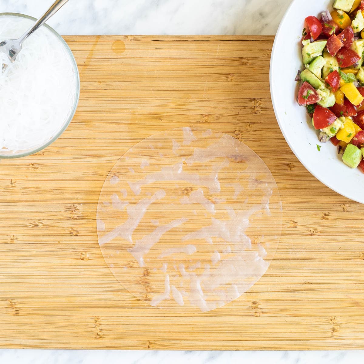 A cutting board with wet rice paper wrapper. A white bowl of colorful chopped veggies and fruits and a small bowl with rice noodles are next to it.