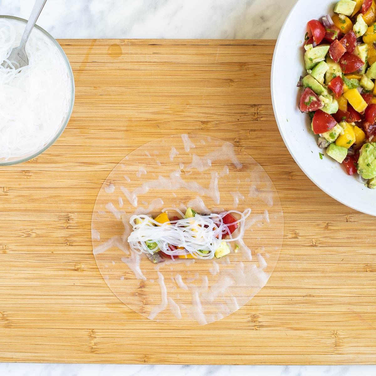 A cutting board with wet rice paper wrapper topped with a small heap of colorful chopped veggies and fruits and rice noodles. More fillings are in separate bowls next to it.