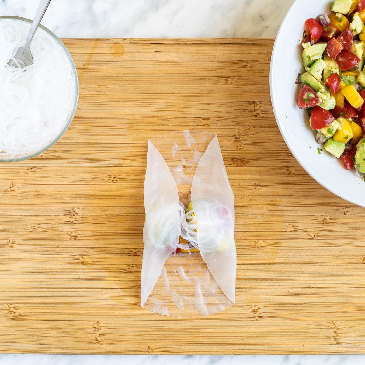 A cutting board with wet rice paper wrapper topped with a small heap of colorful chopped veggies and fruits and rice noodles. The sides are folded to the middle. More fillings are in separate bowls next to it.