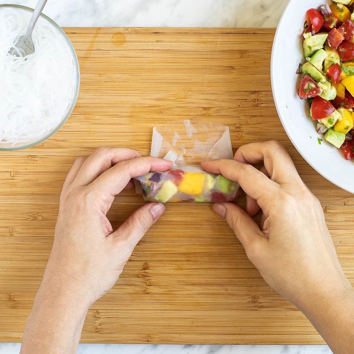 A hand is rolling a wet rice paper wrapper filled with colorful chopped veggies and fruits and rice noodles on a cutting board. More fillings are in separate bowls next to it.