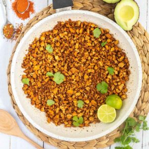 A white frying pan with brown crispy tofu crumbles sprinkled with freshly chopped cilantro. Lime slices are added. Avocado and more spices are next to the pan.