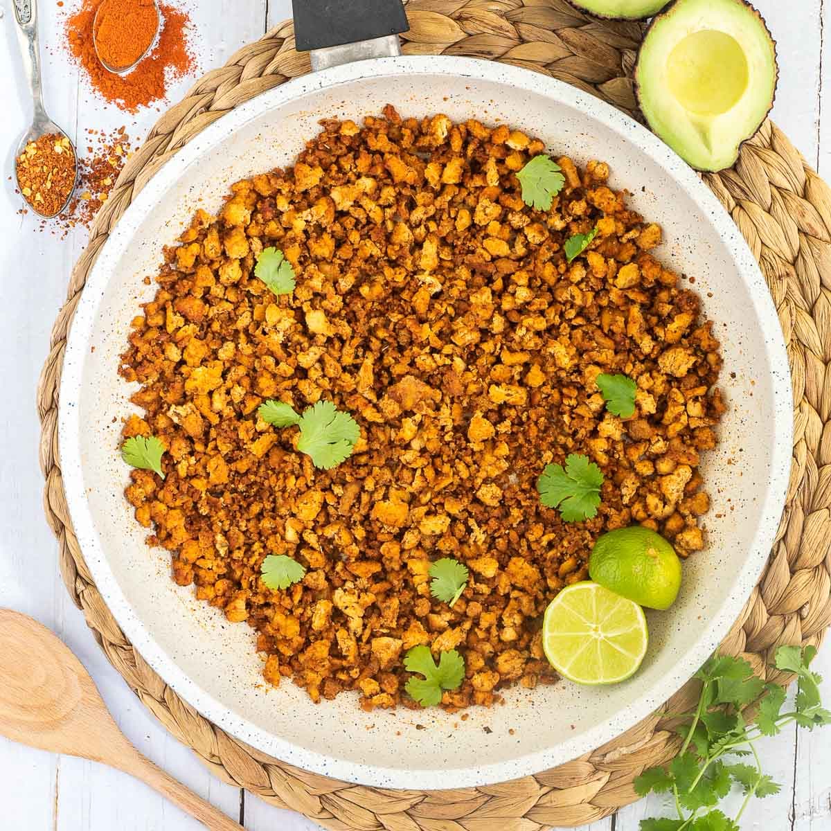 A white frying pan with brown crispy tofu crumbles sprinkled with freshly chopped cilantro. Lime slices are added. Avocado and more spices are next to the pan.