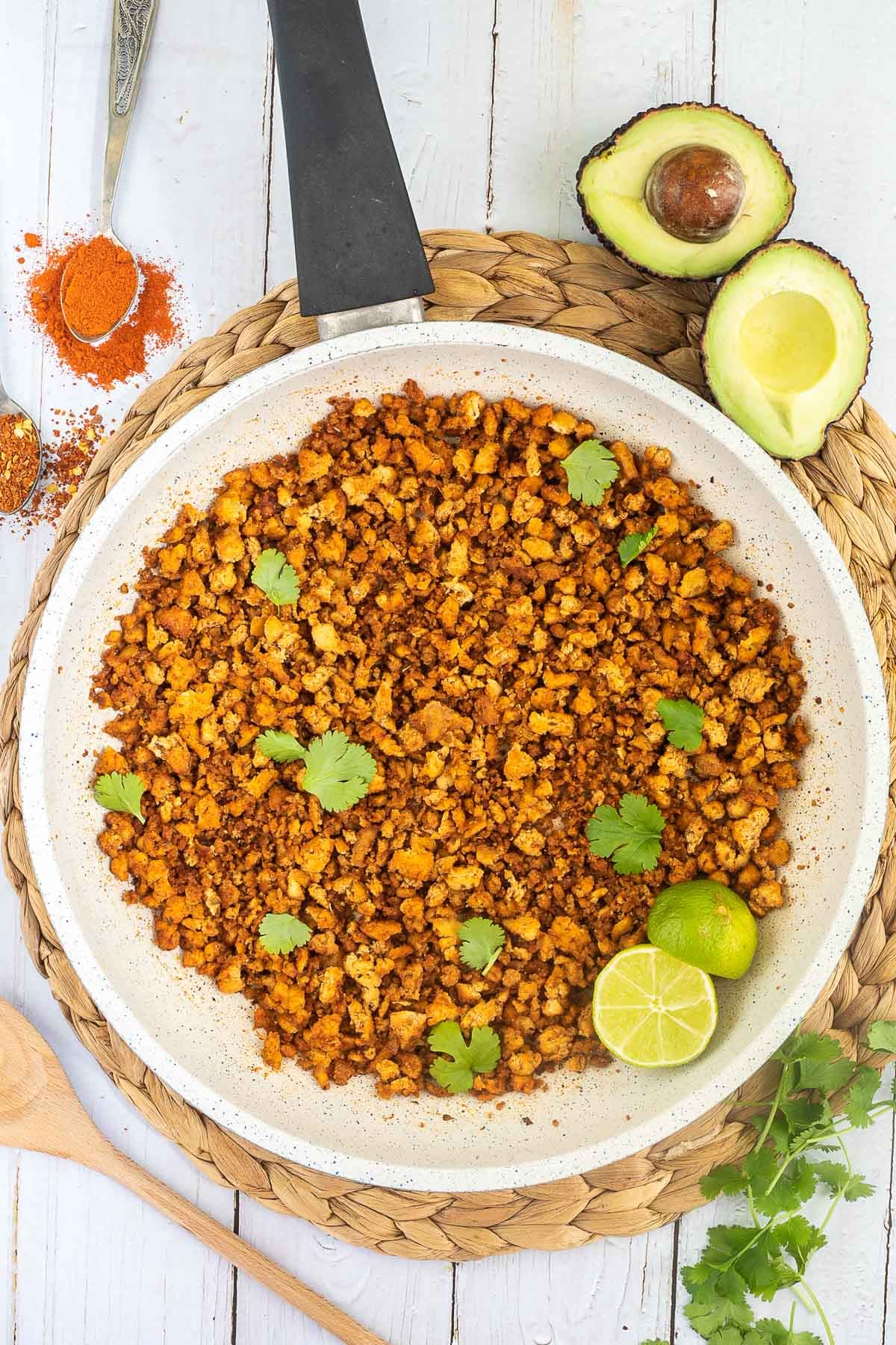 A white frying pan with brown crispy tofu crumbles sprinkled with freshly chopped cilantro. Lime slices are added. Avocado and more spices are next to the pan.