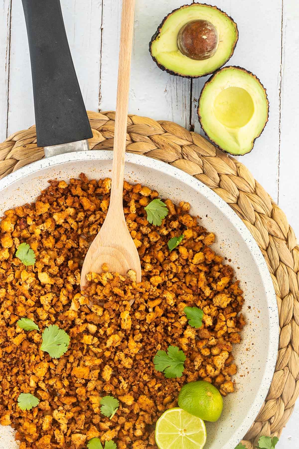 A white frying pan with brown crispy tofu crumbles sprinkled with freshly chopped cilantro. Lime slices and a wooden spatula are added. Avocado and more spices are next to the pan.