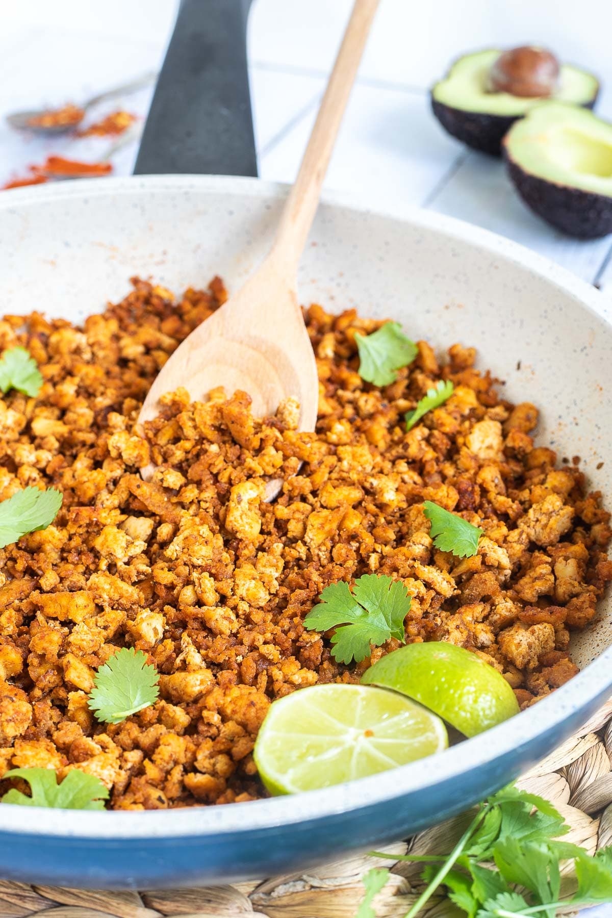 A white frying pan with brown crispy tofu crumbles sprinkled with freshly chopped cilantro. Lime slices and a wooden spatula are added. Avocado and more spices are next to the pan.