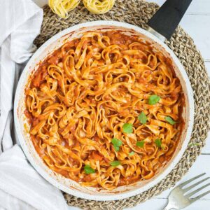 Tagliatelle is mixed in and covered with a creamy light red tomato-based sauce topped with fresh green herbs in a white frying pan. Dry tagliatelle pasta and a cheese grater with parmesan cheese is next to it.