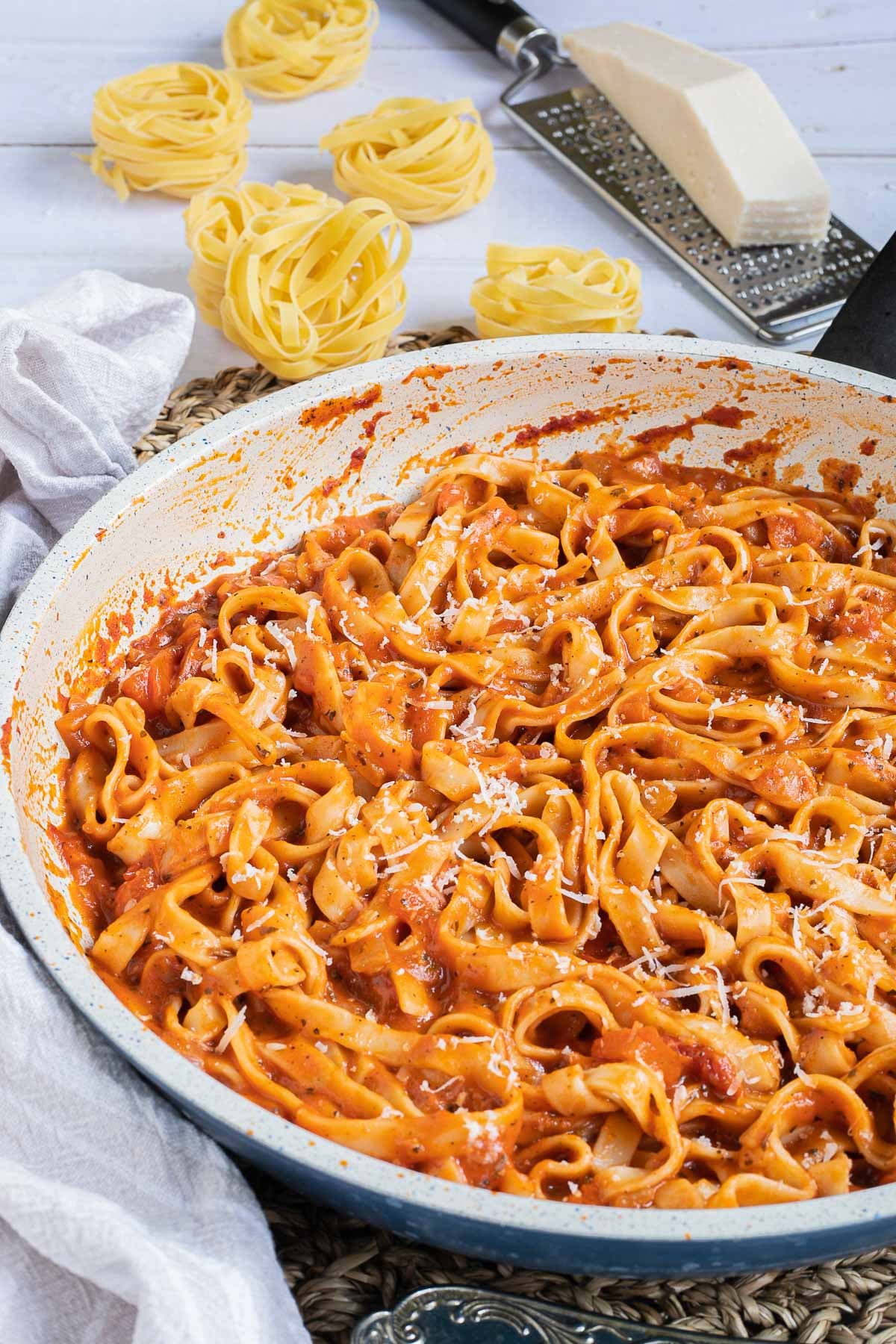 Tagliatelle is mixed in and covered with a creamy light red tomato-based sauce in a white frying pan. Dry tagliatelle pasta and a cheese grater with parmesan cheese is next to it.