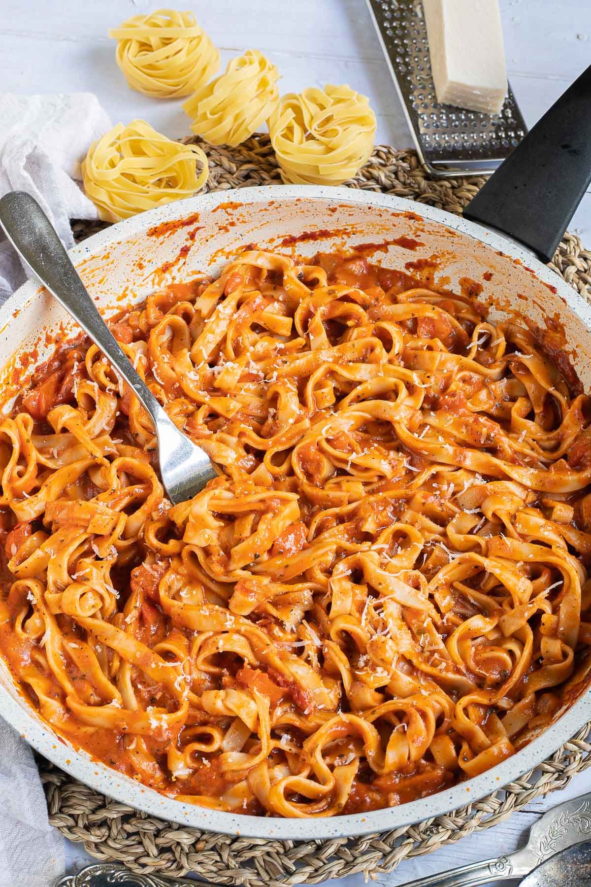 Tagliatelle is mixed in and covered with a creamy light red tomato-based sauce in a white frying pan. Dry tagliatelle pasta and a cheese grater with parmesan cheese is next to it. A fork is dipped in the middle.