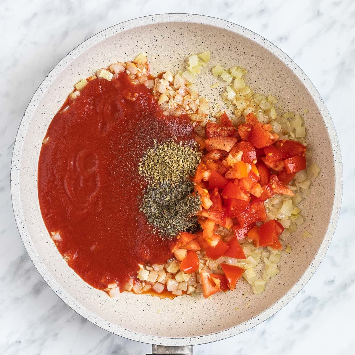 Chopped onion, garlic, tomatoes and tomato sauce with dried herbs in a white frying pan.