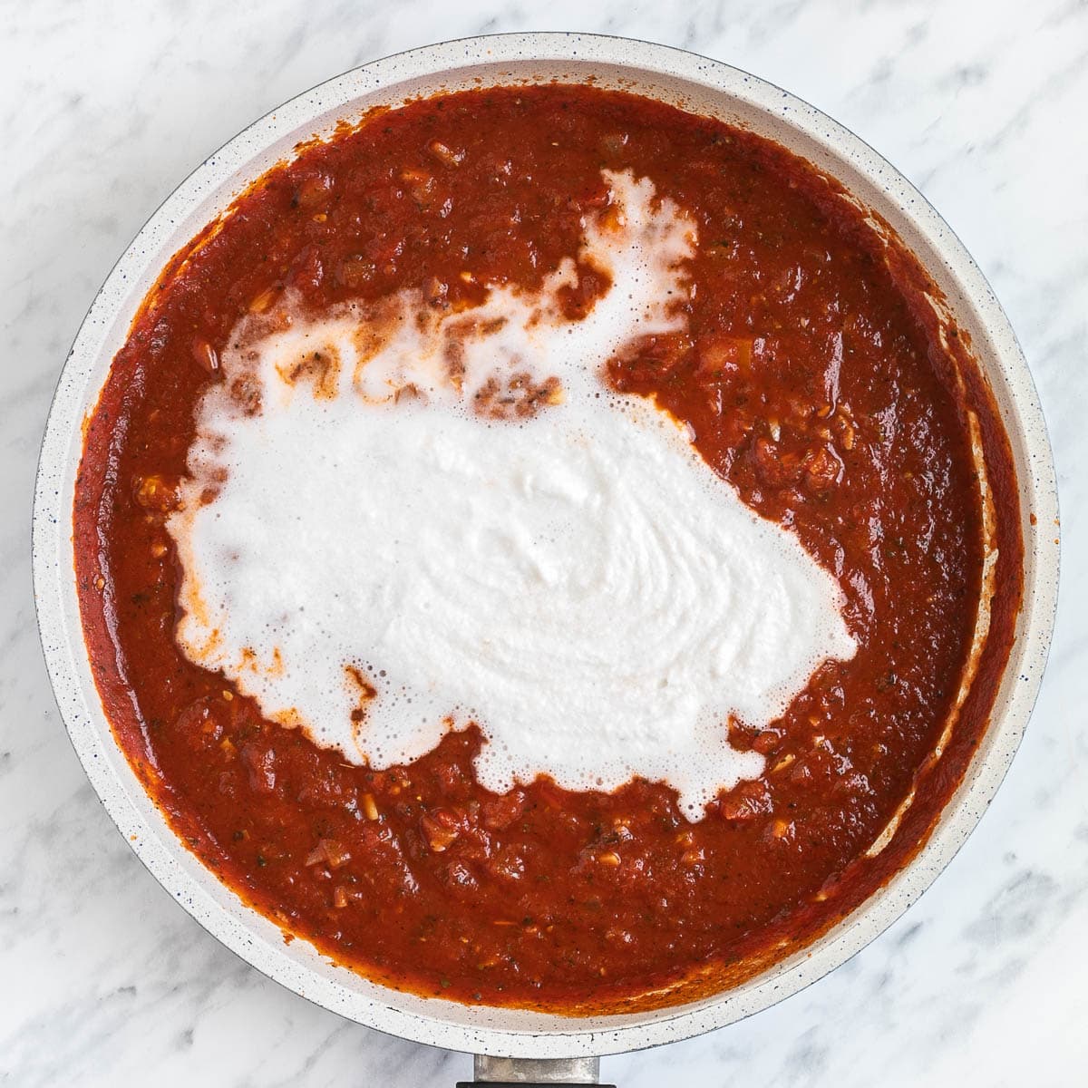 White sauce is in the middle of chunky red tomato sauce in a white frying pan.