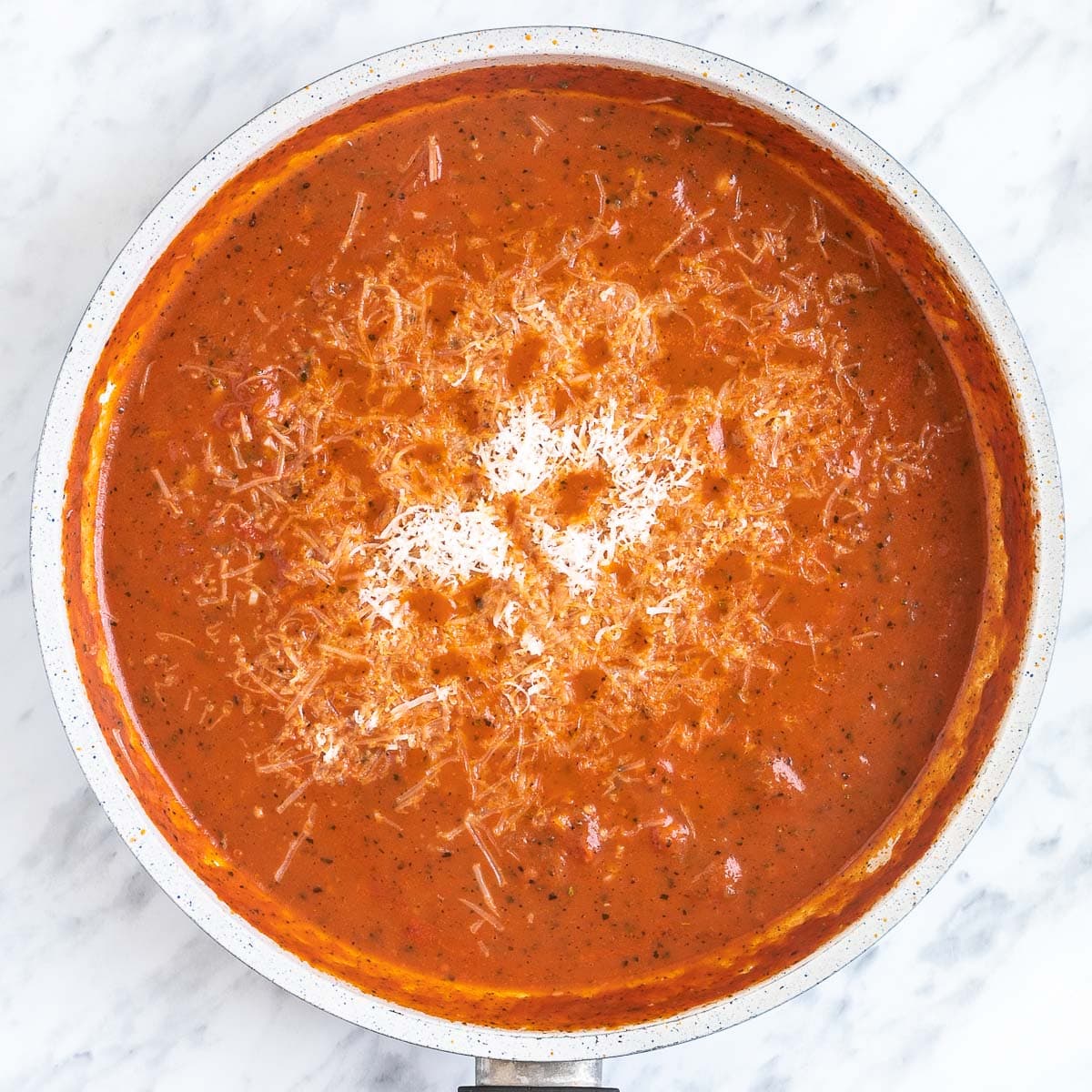 Shredded cheese is added on top of a light red chunky tomato sauce in a white frying pan.