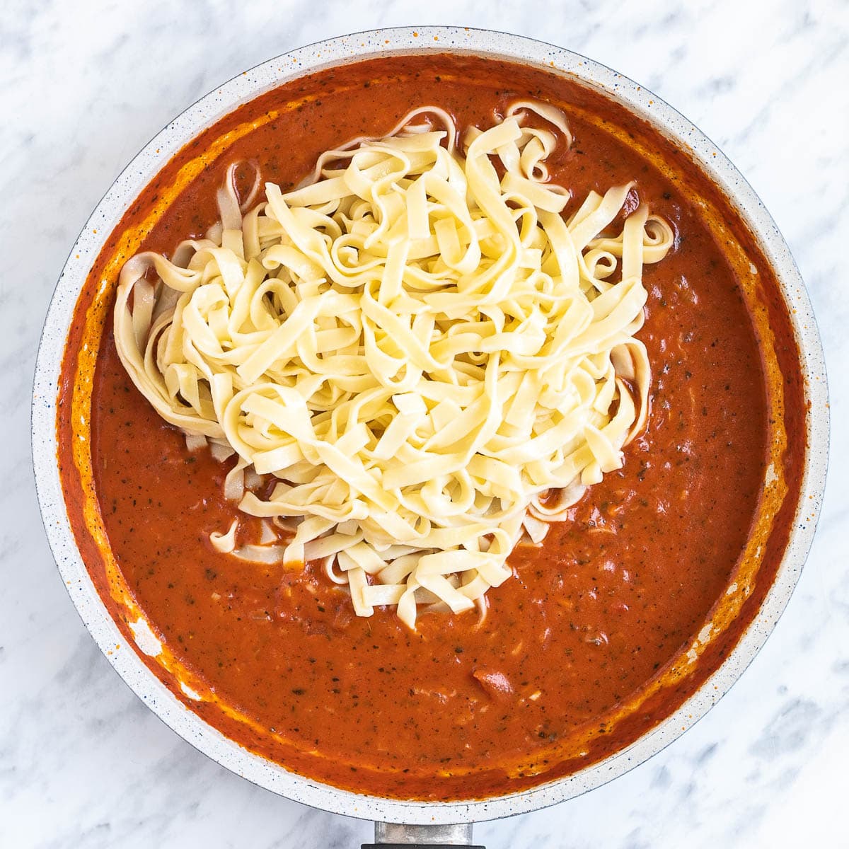 Tagliatelle is added to a light red chunky tomato sauce in a white frying pan.