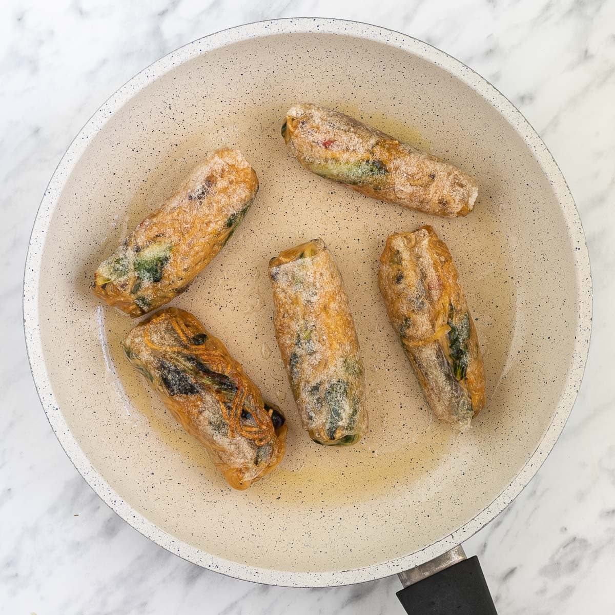 White frying pan with 5 ramen spring rolls that are getting crispy and shiny.