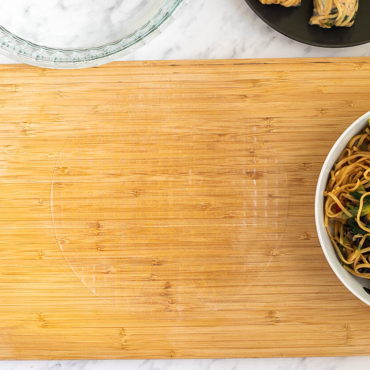 A wooden cutting board with wet rice paper. Ramen noodles in a white bowl. Water in a glass bowl and 2 finish spring rolls on a black plate around it.