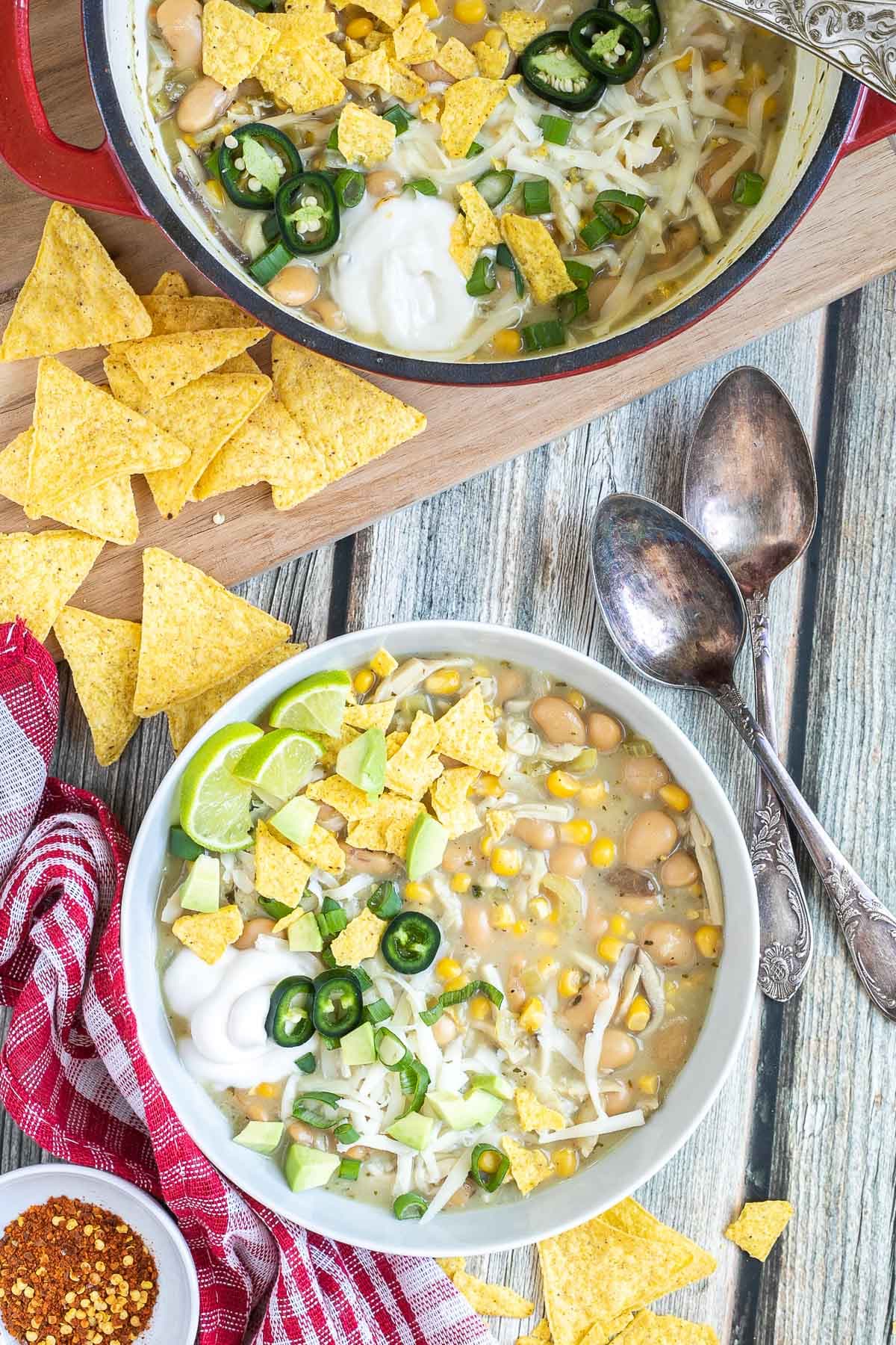 A white bowl with a light brown chili with white beans, shredded mushrooms, corn, topped with green chili slices, shredded cheese and nacho chip pieces. Leftover chili in the pot is right next to it.
