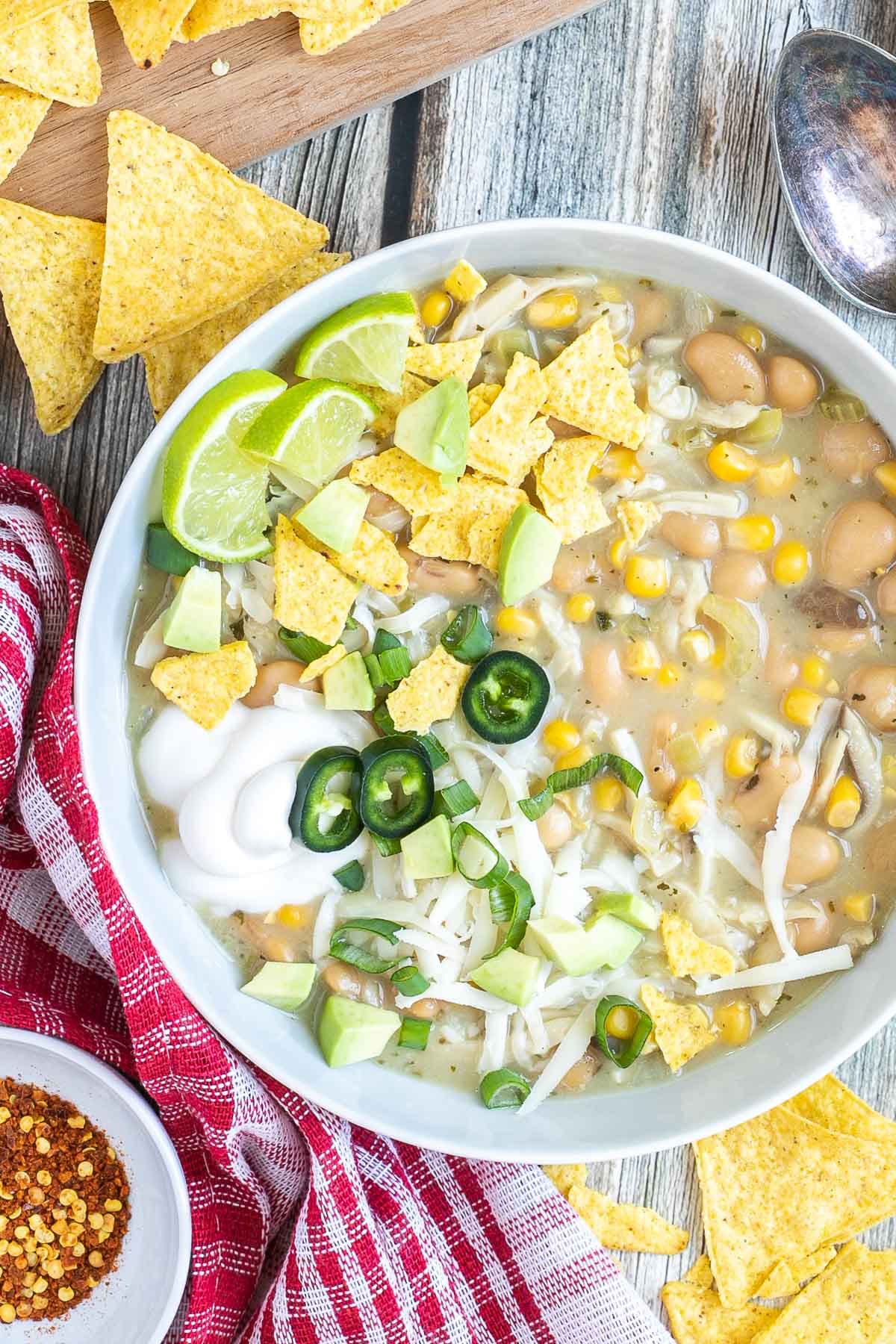 A white bowl with a light brown chili with white beans, shredded mushrooms, corn, topped with green chili slices, shredded cheese and nacho chip pieces.