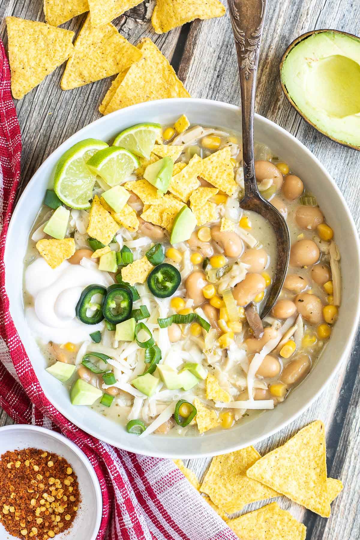A white bowl with a light brown chili with white beans, shredded mushrooms, corn, topped with green chili slices, shredded cheese and nacho chip pieces.
