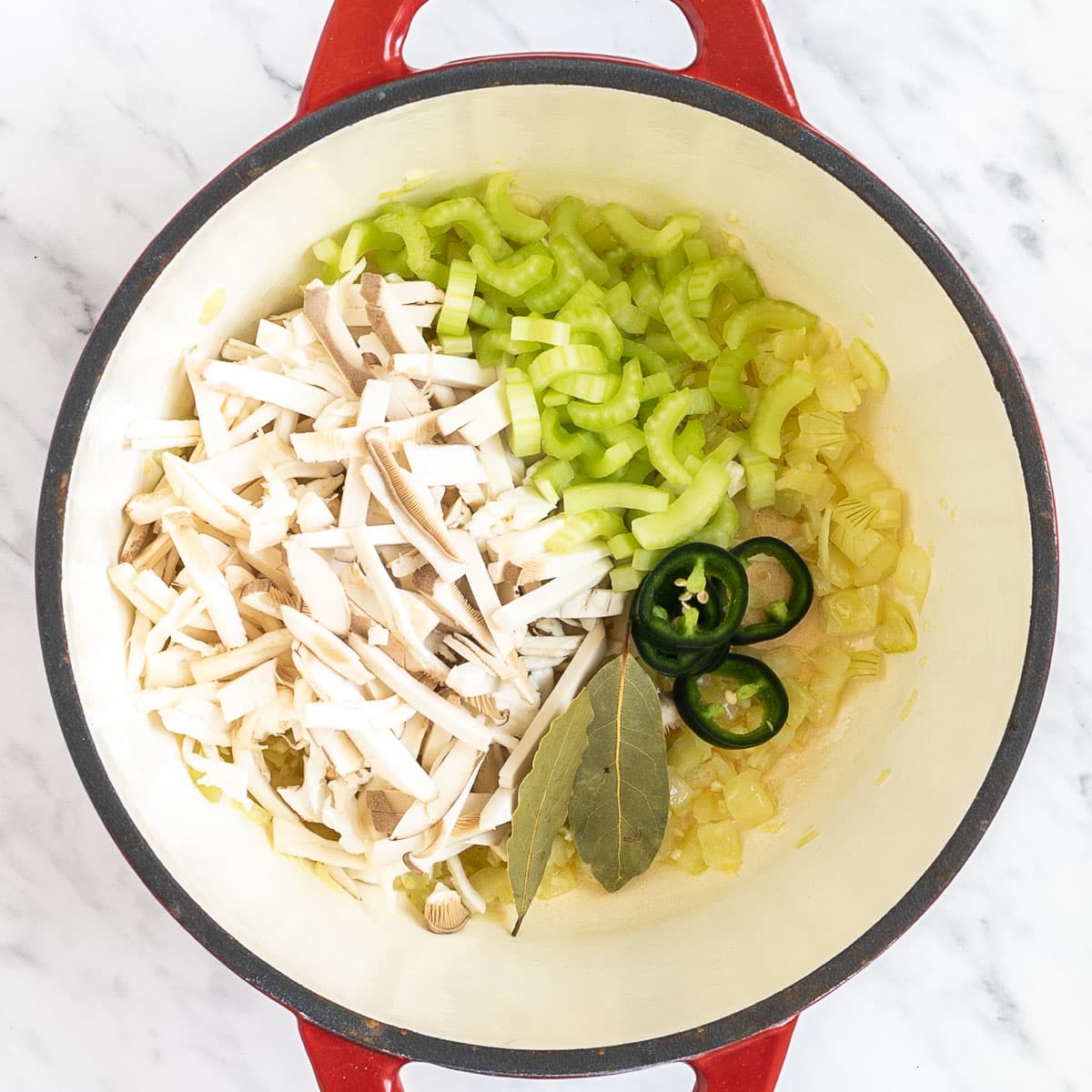 Red Dutch oven with shredded mushrooms, celery slices, chopped onion, bay leaves, and red chili pepper slices.