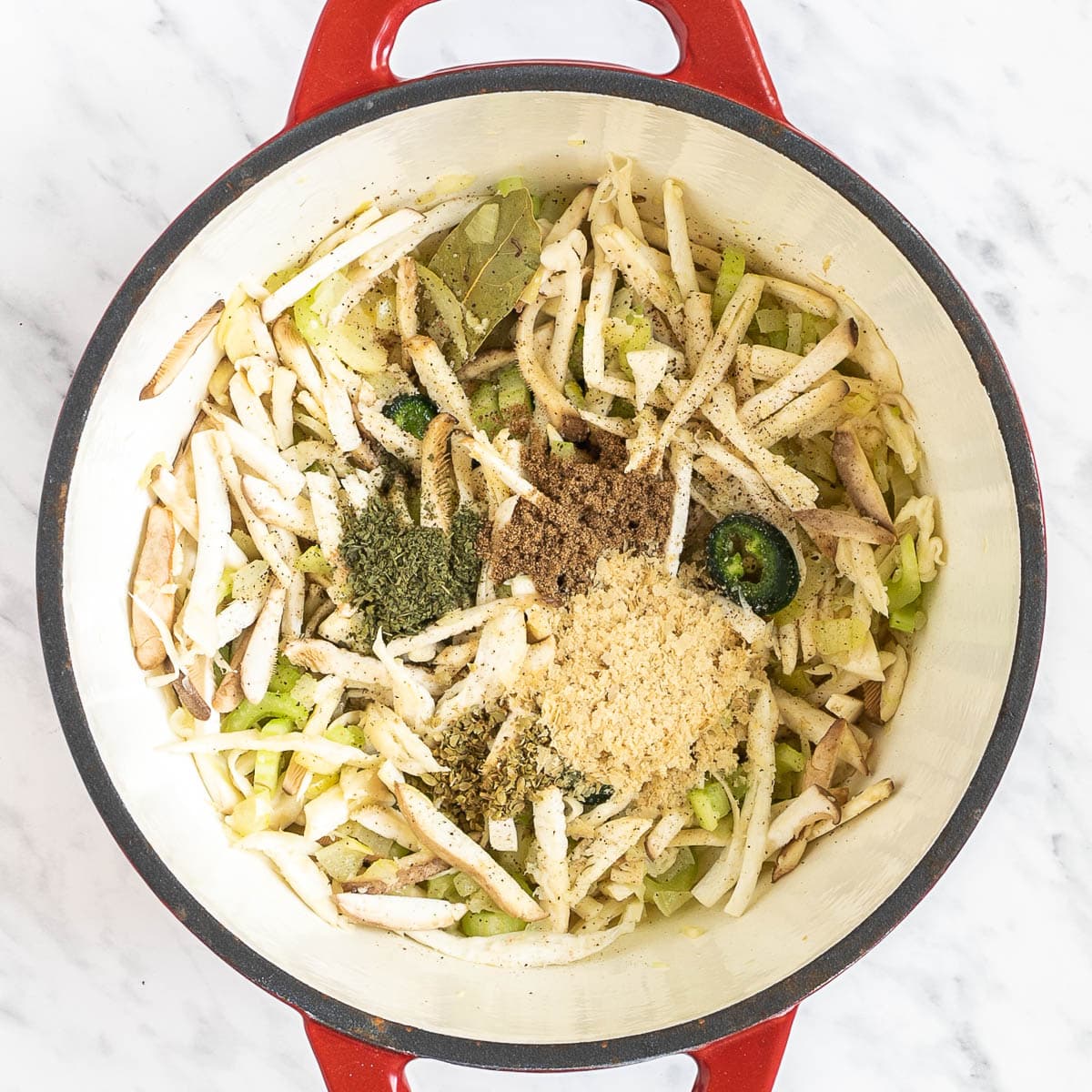 Red Dutch oven with shredded mushrooms, celery slices, chopped onion, bay leaves, and red chili pepper slices mixed together and topped with heaps of different spices.
