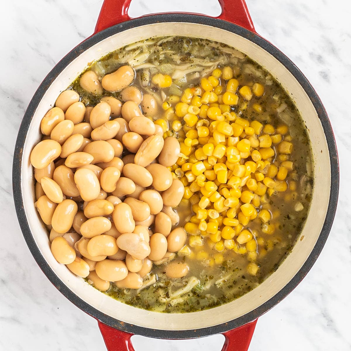 Red Dutch oven with shredded mushrooms in a thin light green white soup topped with white beans and sweet corn.